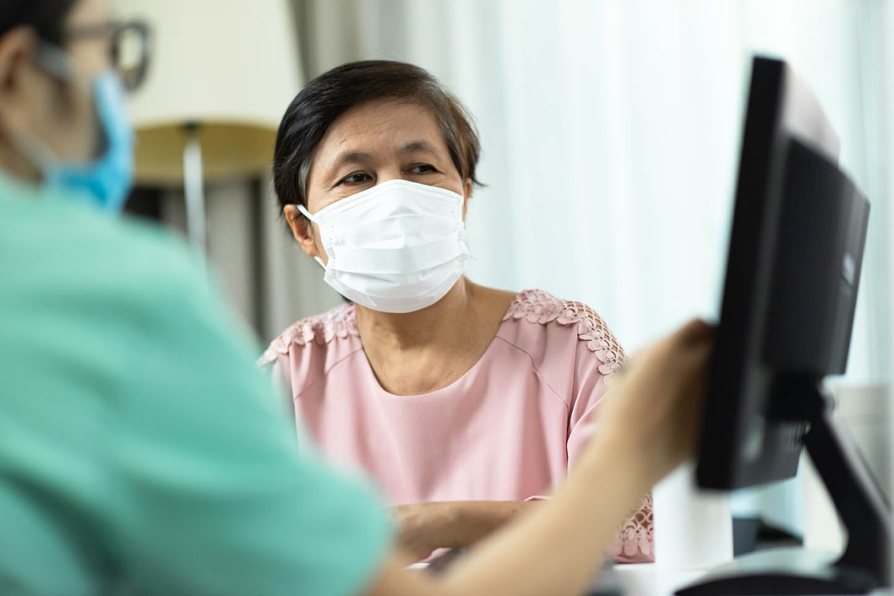Asian Female Doctor or Nurse in green uniform talking giving advice to Elderly woman patient in pink cloth and looking to computer screen monitor together in medical room at the hospital. Disease, Ill, Flu, Sick of people. Covid-19, Coronavirus.